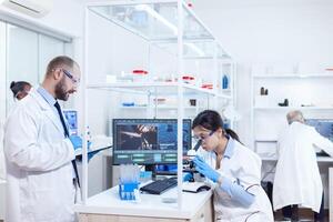 Male scientist taking notes on tablet pc during virus study. Team of researchers doing pharmacology engineering in sterile laboratory for healthcare industry with african assistant in the background. photo