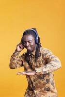 Smiling cheerful man enjoying song on headphones in studio over yellow background, dancing and having fun during leisure time. African american person relaxing and listening to audio on camera photo