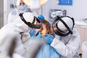 Dentist woman treating little girl bad tooth using drill with help from assistant dressed in coverall. Stomatology team wearing ppe suit during covid19 doing procedure on child teeth. photo