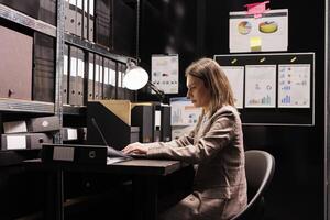 Woman manager typing administration report on laptop computer, working overtime at management research in file room. Bookkeeper checking accountancy record, searching for business documentation photo