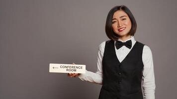 Asian receptionist pointing at wall indicator for conference room, using pointer to help customers find all amenities and facilities. Woman employee holding sign to indicate directions. Camera B. photo