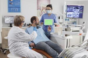 Assistant woman holding mock up green screen chroma key tablet with isolated display discussing oral hygiene with man patient during stomatological consultation in dental office. stomatology concept photo