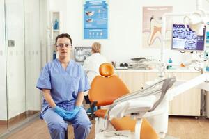 Closeup of medical assistant sitting on chair in hospital office waiting for sick man patient to examining dental problem during stomatological appointment. In background senior doctor checking x-ray photo