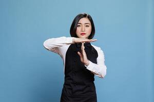 Confident asian waitress dressed in restaurant uniform making timeout gesture with arms portrait. Woman receptionist showing take a break sign with hands and looking at camera with serious expression photo