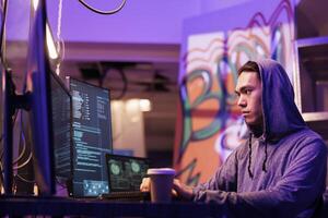 Asian hacker stealing information from server using computer worm. Criminal in hood coding illegal internet virus and breaking into server while drinking coffee to go in dark room photo