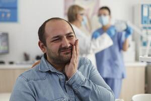 Close up of man patient with toothache sitting on dental chair during stomatological examination waiting for medical expertise. In background specialist team discussing dentistry treatment photo