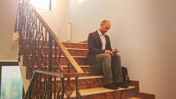 Businessman sitting alone on stairs typing on smartphone in finance corporate company working overtime. Group of professional successful businesspeople working in modern financial building. photo