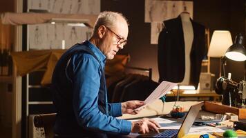 Senior couturier work on items design with sketches, using laptop to review material and custom made sizes. Male professional tailor creating luxury clothes in workshop atelier. photo