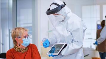 Woman sitting on chair in waiting area with protection mask listening doctor with overall looking on tablet in clinic with new normal. Assistant explaining dental problem during coronavirus pandemic photo