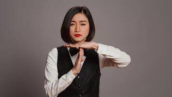Asian receptionist expressing timeout symbol on camera, showing negativity and asking for a work break. Tired bored employee showing pause sign in studio over grey background. Camera B. photo