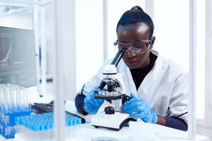 African chemist doing investigation using microscope analysing sample on glass slide. Black healthcare scientist in biochemistry laboratory wearing sterile equipment. photo