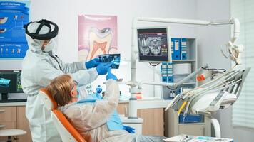 Dentist with face shield reviewing panoramic mouth x-ray image to patient during global pandemic. Assistant and doctor talking with senior woman wearing suit, coverall, protection suit, mask, gloves photo