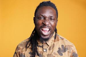 Furious stressed man having breakdown while posing aggresive in studio over yellow background, showing unpleasant reaction. African american young adult feeling frustrated and mad photo