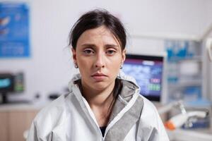 Portrait of tired dentist in protective gear agasint coronavirus looking at camera. Exhausted medical specialist in dentistry cabinet wearing safety equipment during global health crisis. photo