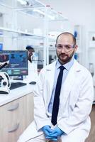 Healthcare researcher wearing lab coat and protective glasses looking at camera. Serious expert in genetics in lab with modern technology for medical investigations with african assistant in the background. photo