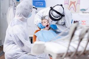 Little girl with mouth open getting caries treatment from pediatric dentist dressed in ppe suit. Dentist in coronavirus suit using curved mirror during teeth examination of child. photo