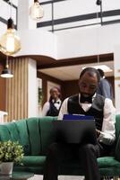 Computer and hotel industry. Black guy bellboy sitting with laptop in lobby entering data filling online document. African American man concierge making entry in daily luggage movement register photo