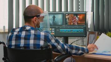 Disabled man videographer with face protection editing video project creating content sitting in wheelchair in new normal office. Blogger working durig global pandemic respecting social distance. photo