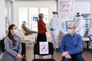 Senior woman patient in stomatology clinic discussing with doctor dressed in ppe suit as safety precaution during global pandemic with coronavirus. People talking in stomatology waiting room. photo