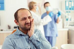 Man patient complaining about teeth pain while waiting for stomatologist doctors to check toothache. Senior orthodontic doctor and medical nurse examining x-ray working in hospital stomatology clinic photo