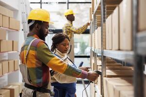 Warehouse manager supervising assistant scanning parcel barcode and pointing at cardboard box on shelf. African american storehouse worker using scanner on package and working with supervisor photo