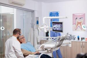 Senior woman discussing with dentist in dental cabinet about teeth issue sitting on chair. Medical teeth care taker discussing with senior woman about mouth hygiene. photo