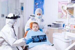 Dentist in coverall exaplaining dental procedure to kid and parent dressed in ppe suit showing radiography. Stomatolog in protectie suit for coroanvirus as safety precaution holding child teeth x-ray during consultation. photo