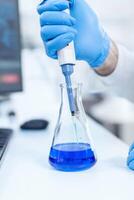 Close up of scientist testing liquid in laboratory flask using micro pipette. Senior professional chemist using pippete with blue solution for microbiology tests. photo