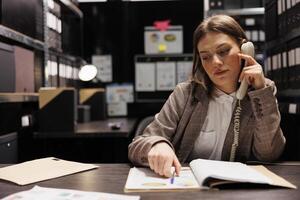 Bookkeeper having remote conversation, working overtime at administrative report in storage room. Caucasian businesswoman discussing accountancy folders, checking corporate documentation photo