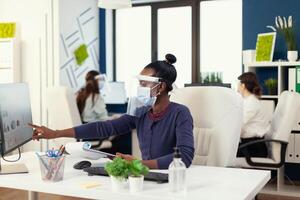 African marketing specialist at workplace wearing face mask during coronavirus. Multiethnic coworkers working respecting social distance in financial company. photo