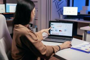Busy employee pointing at graph working to finish a deadline wprlomg late. Businesswoman looking at statistics on laptpop screen sitting at her workplace doing overtime. photo