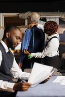 Teamworking tailors helping senior client find perfectly fitting bespoke business suit. Cheerful couturiers taking elderly gentleman measurements during sartorial customization comission process photo