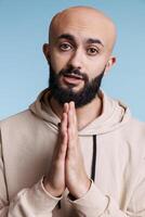 Young adult arab man with folded arms asking for help while looking at camera with pleading facial expression. Person pleasing, praying to god and making wish studio portrait photo