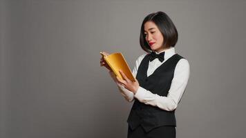 Asian receptionist reading a literature book over grey background, enjoying lecture hobby with a novel story in studio. Hotel concierge employee reads a fiction tale for knowledge. Camera A. photo