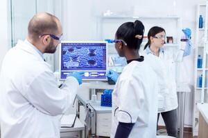 African doctor and scientist working conducting virus experiment using computer software. Multiethnic team of medical researchers working together in sterile lab wearing protection glasses and gloves. photo