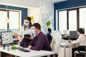 international team doing financial analysis wearing face mask as safety precaution during global pandemic. Multiethnic team in new normal financial office in corporate company typing on computer, taking notes on tablet. photo