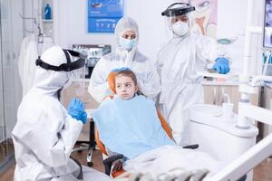 Dentist explaining dental problem dressed in protection suit in the course of coronavirus pandemic. Stomatologist during covid19 wearing ppe suit doing teeth procedure of child sitting on chair photo