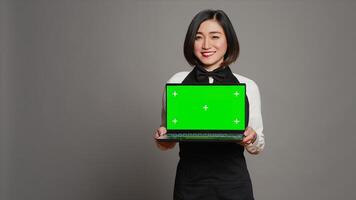 Asian server presenting greenscreen display on laptop in studio, wearing uniform with apron and bow. Restaurant hostess holding pc with blank mockup template, copyspace layout. Camera A. photo