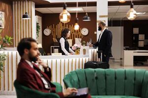 Businessman chats with receptionist in hotel lobby, expressing gratitude to front desk staff for assistance. White collar worker appreciating easy check in experience on business trip. photo