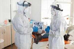 Dentist doctors with ppe suit analysing teeth x-ray using tablet in dental room, planning surgery during global pandemic while patient waiting on stomatological chair. photo