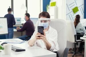 Businesswoman chatting on smartphone sitting at her workplace in busy office wearing face mask during global pandemic with covid19. Multiethnic coworkers working respecting social distance in financial company. photo