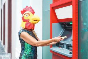 Mature woman wearing cock mask withdraw money from bank cash machine with debit card - Surreal image of half human and animal - Absurd and crazy concept of ATM advertise photo