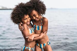 Happy sisters embracing inside sea water during summer time - Afro kids having fun playing on the beach - Family love and travel vacation lifestyle concept photo