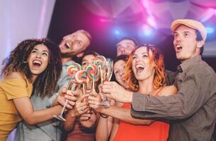 Happy friends having fun toasting with champagne and eating lollipops in disco club - Group young people celebrating together new year party - Youth culture holidays lifestyle concept photo