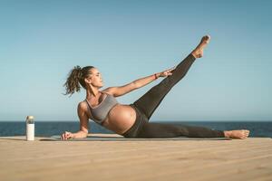 Young pregnant woman doing prenatal pilates exercises session next the sea - Health lifestyle and maternity concept photo