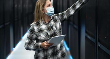 Female data center technician working inside server rack room while wearing face mask photo