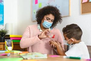 Teacher cleaning hands to stundet children with sanitizer gel while wearing face mask in preschool classroom during corona virus pandemic - Healthcare and education concept photo