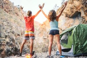 Health young couple doing yoga next to fire while camping with tent on a mountain - Friends meditating together spreading their hands up - People, travel lifestyle and healthy sports concept photo