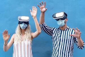 Young couple wearing face surgical mask having fun with virtual reality glasses during outbreak - Millennial people using innovated vr headsets goggles - Youth generation and technology concept photo
