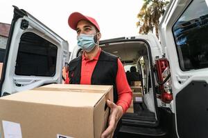 Young delivery man carrying cardboard box while wearing face mask to avoid corona virus spread - People working with fast deliver during corona virus outbreak photo